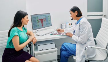 A doctor in a white coat consults with a patient in a teal shirt in a medical office. A computer screen displays gynecological information.