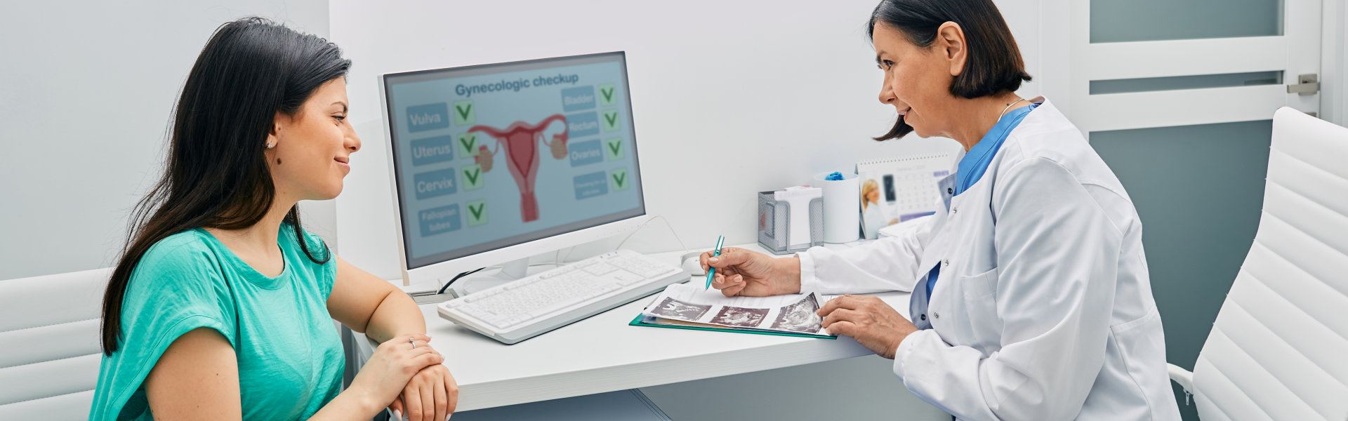 A doctor in a white coat consults with a patient in a teal shirt in a medical office. A computer screen displays gynecological information.